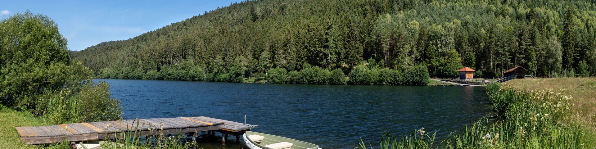 Un lago con un muelle y un bote pequeño, rodeado de un bosque denso y un cielo azul despejado.