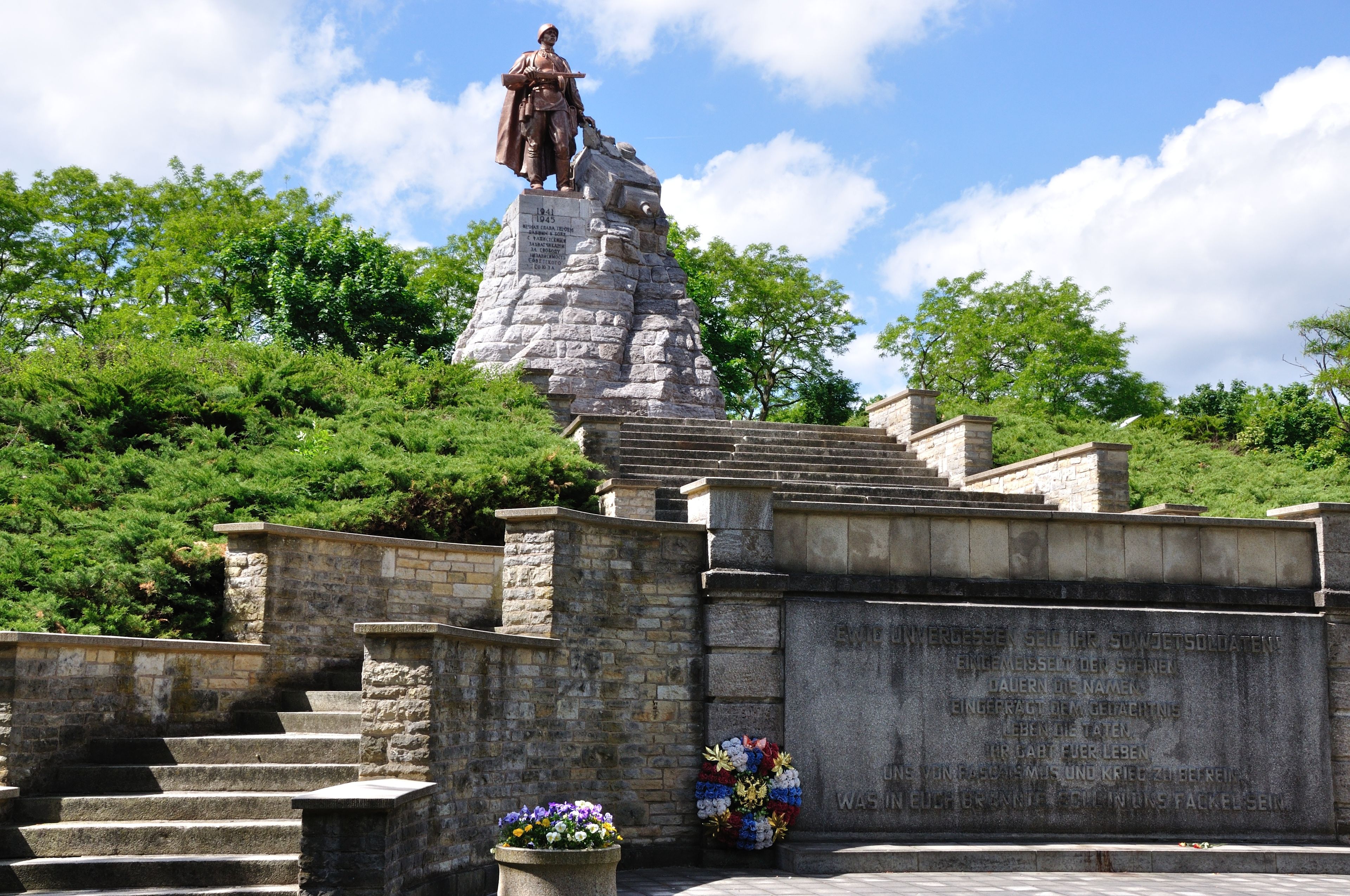 A statue on a stone pedestal with inscriptions, surrounded by stone steps and greenery.