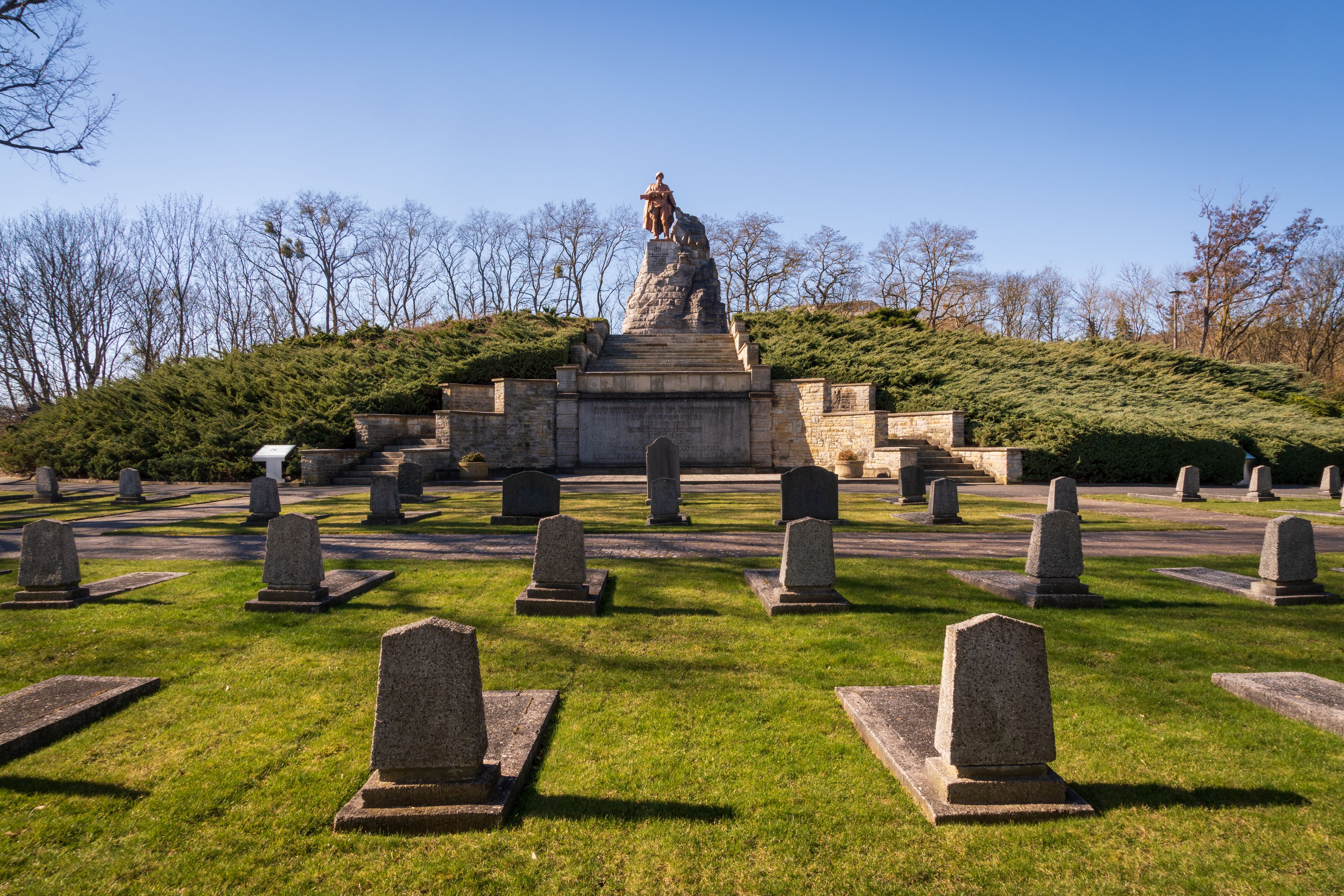 A cemetery with a prominent statue on a hill, surrounded by gravestones and trees.