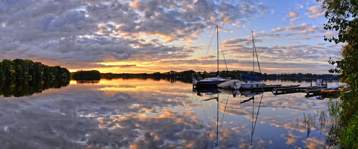 A calm lake with boats docked at a pier, surrounded by trees and a sunset sky.