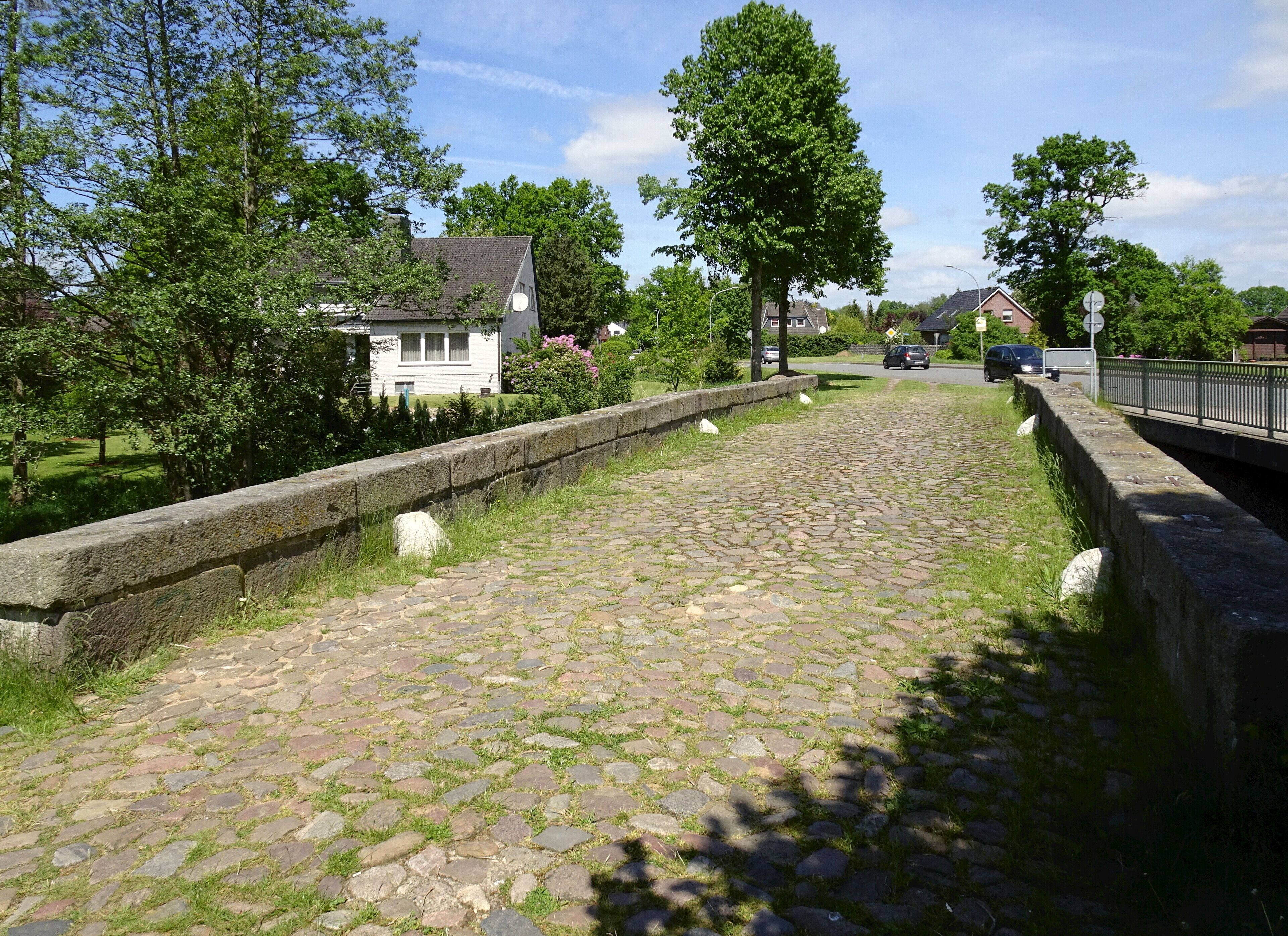 A cobblestone pathway with stone walls, surrounded by greenery and houses.