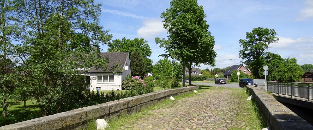 A cobblestone pathway with stone walls, surrounded by greenery and houses.
