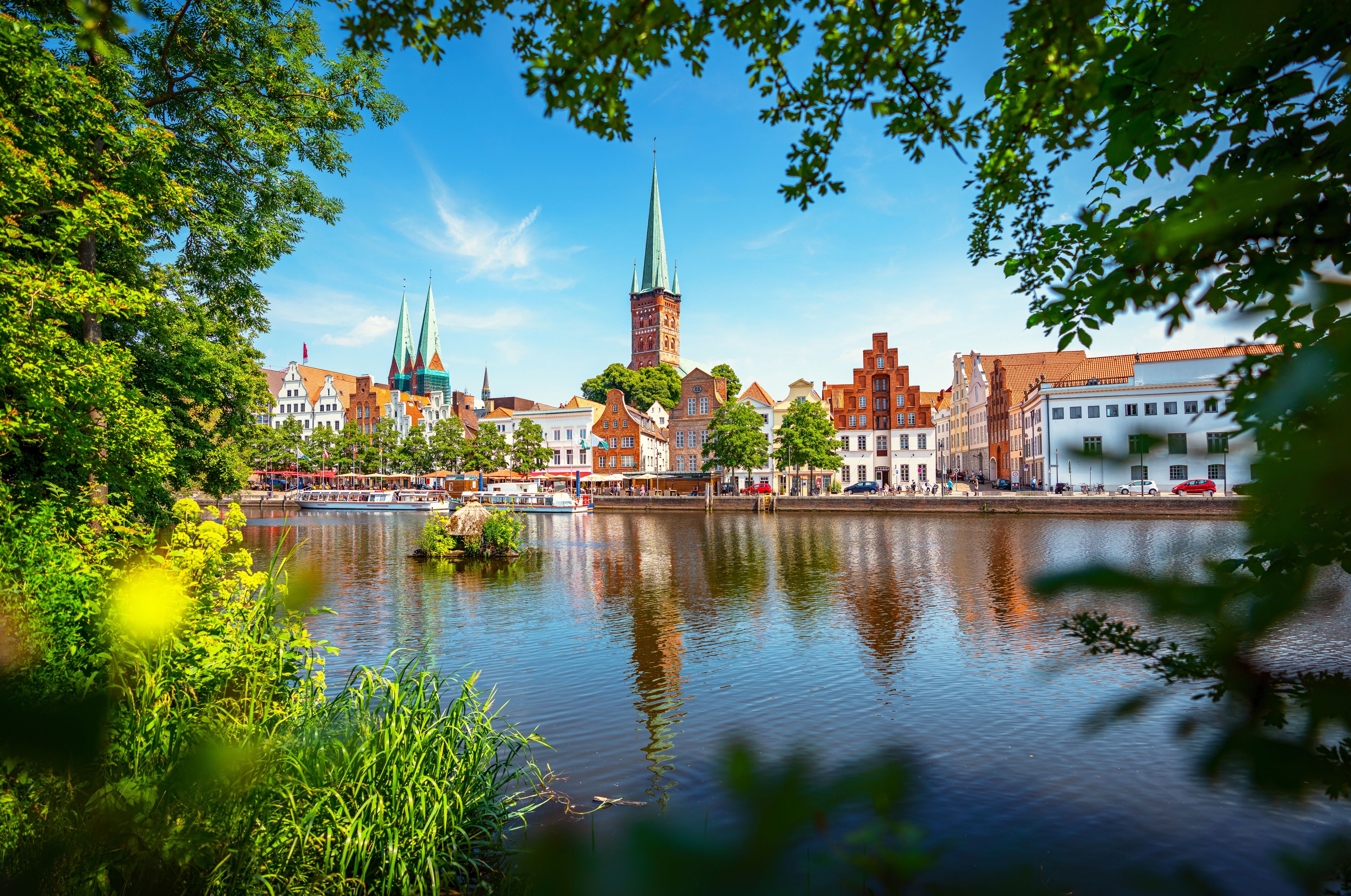 Bybillede ved en flod med historiske bygninger, et markant kirktårn og en rolig flod der spejler arkitekturen.