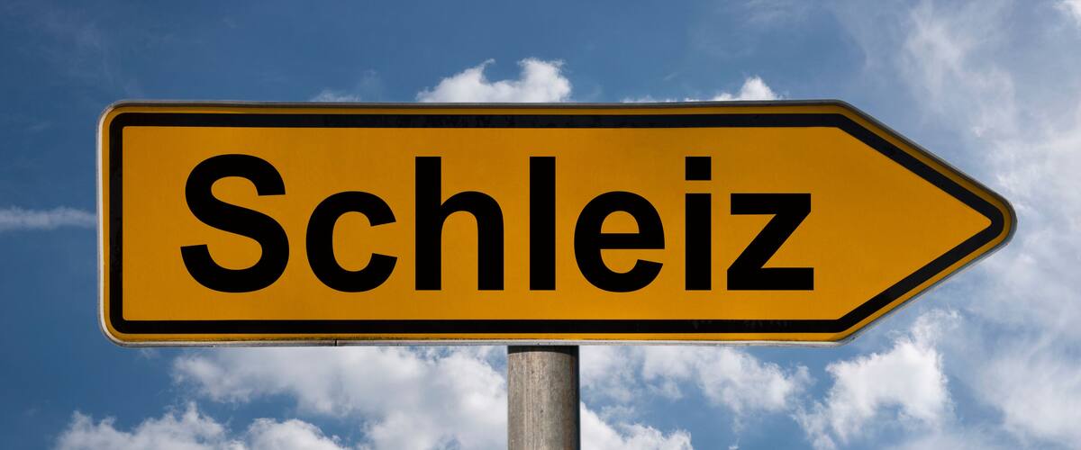 A directional sign with the word "Schleiz" against a blue sky with clouds.