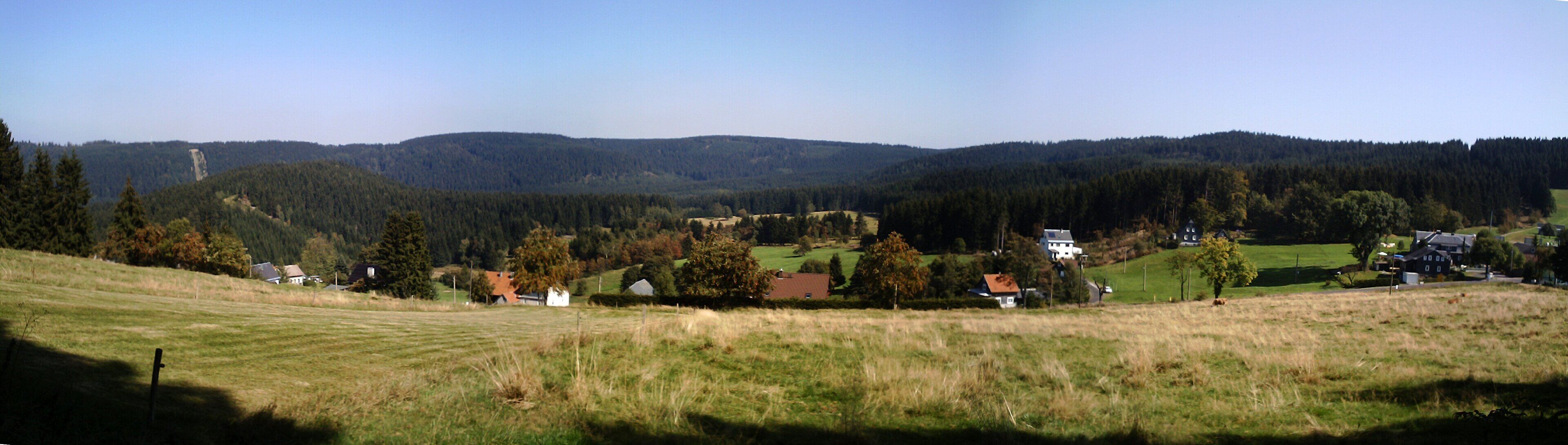 A rural landscape with rolling hills, a forested mountain range, and scattered houses.