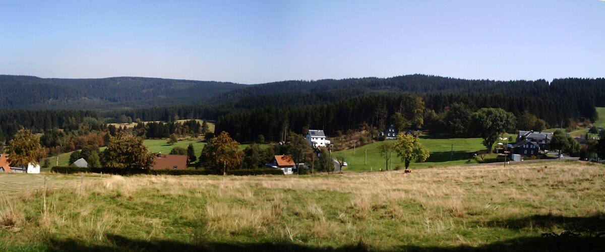 A rural landscape with rolling hills, a forested mountain range, and scattered houses.