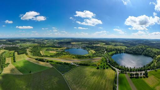 Aerial view of two tranquil lakes nestled amid a tapestry of lush green fields and dense forests under a sky dotted with fluffy clouds, Schalkenmehren, Rheinland-Pfalz, Germany.