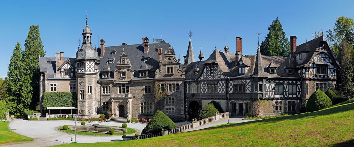 A large, historic building with multiple towers and turrets, surrounded by well-maintained gardens and trees.