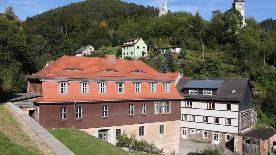 A hillside with a large building with a red roof, a church with a clock tower, and a forested hill in the background.
