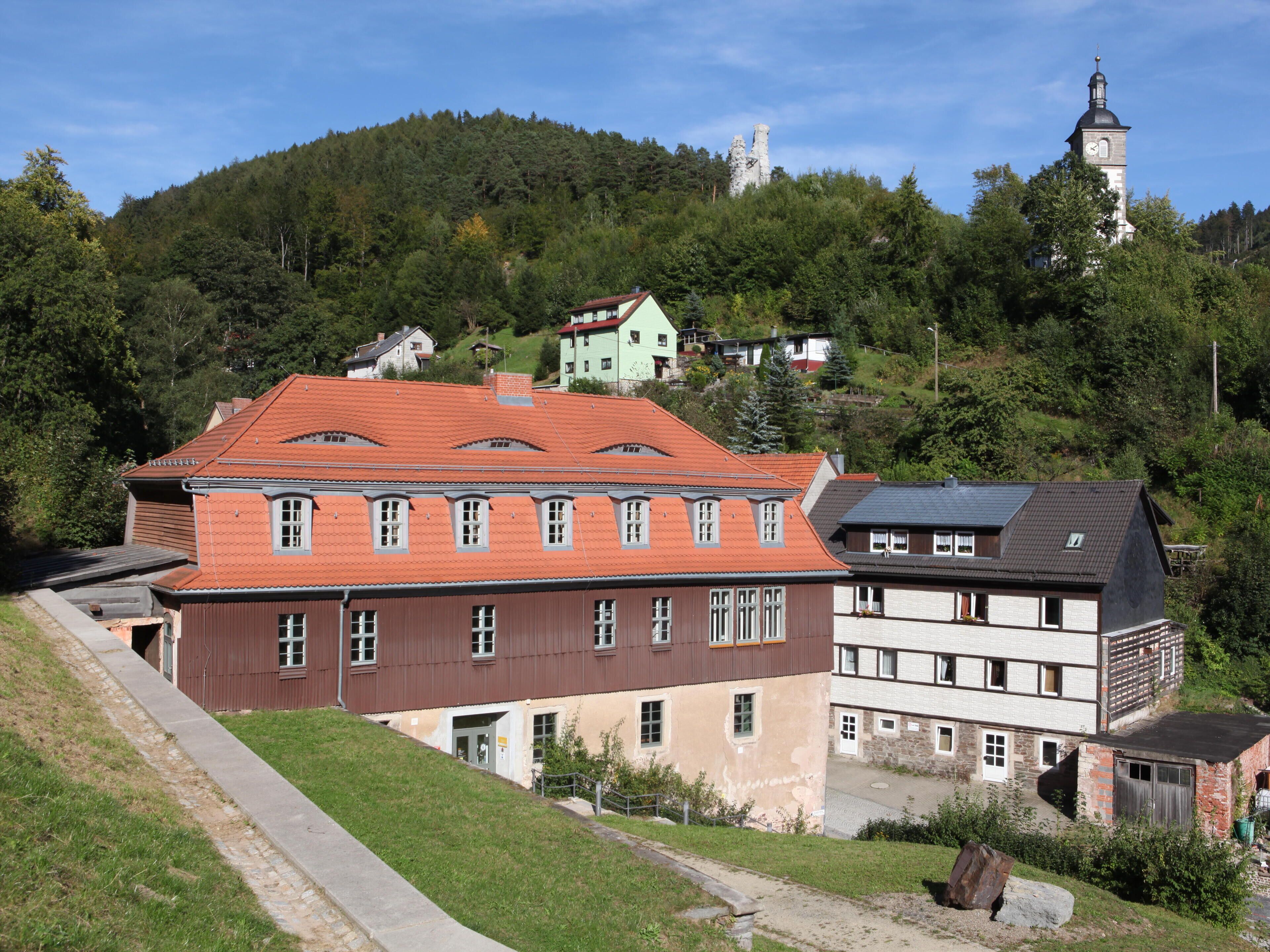 A hillside with a large building with a red roof, a church with a clock tower, and a forested hill in the background.