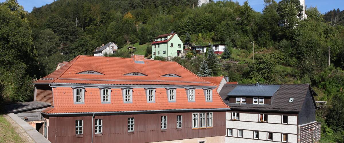 A hillside with a large building with a red roof, a church with a clock tower, and a forested hill in the background.