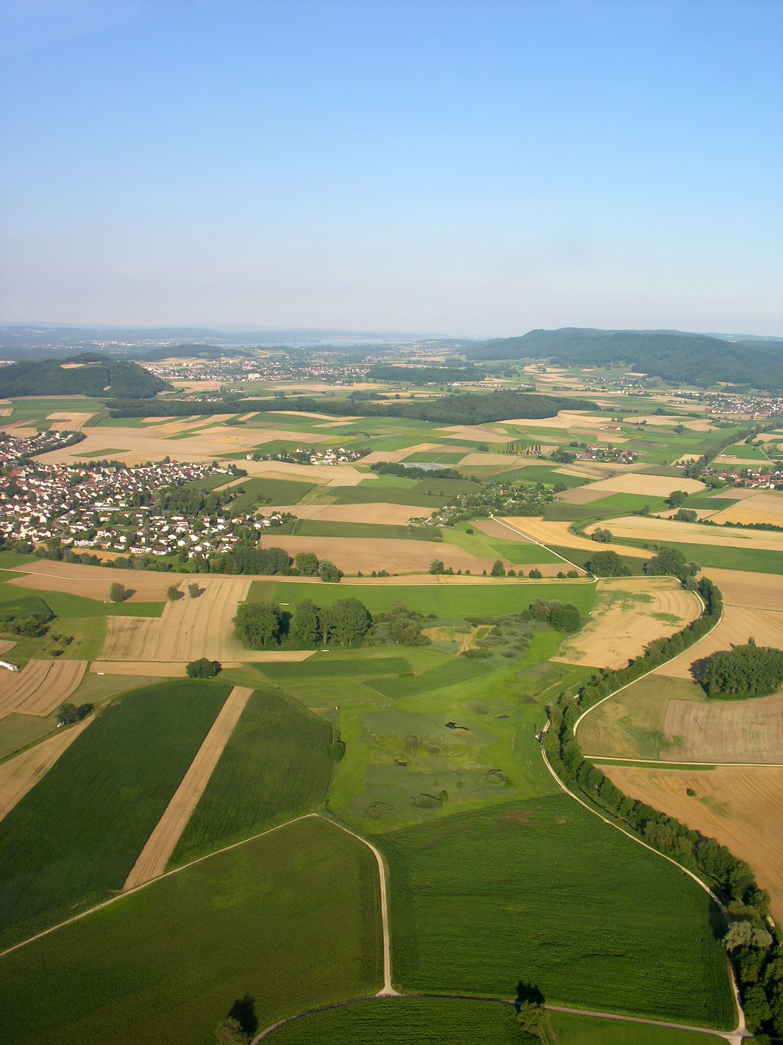 An aerial view of a rural landscape with fields, a winding road, and a small town in the distance.