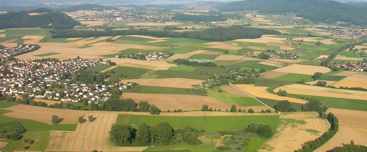 An aerial view of a rural landscape with fields, a winding road, and a small town in the distance.