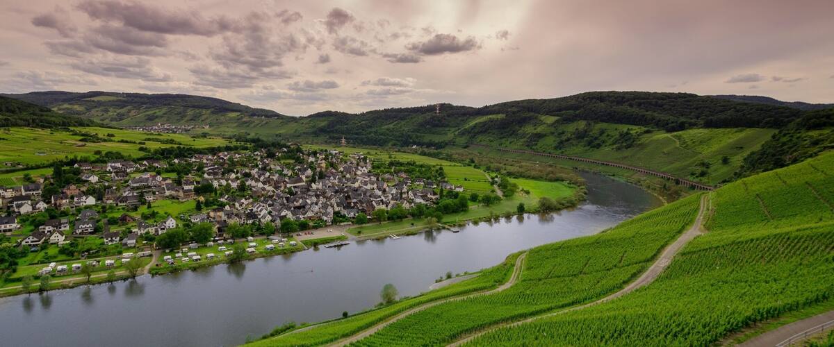 A village by a river with green fields and hills in the background.