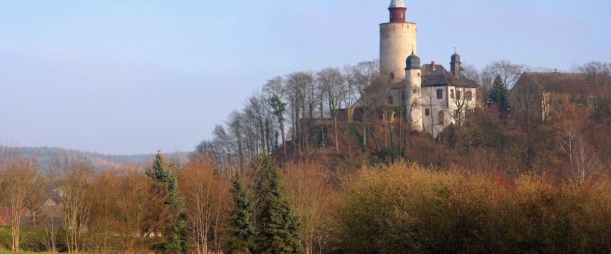 Eine Burg auf einem Hügel mit einem Turm, umgeben von Bäumen und einem grünen Feld.