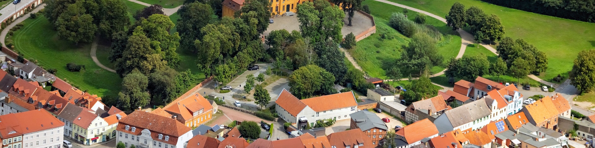 An aerial view of a historic town with red-roofed buildings, a large church, and surrounding green fields.