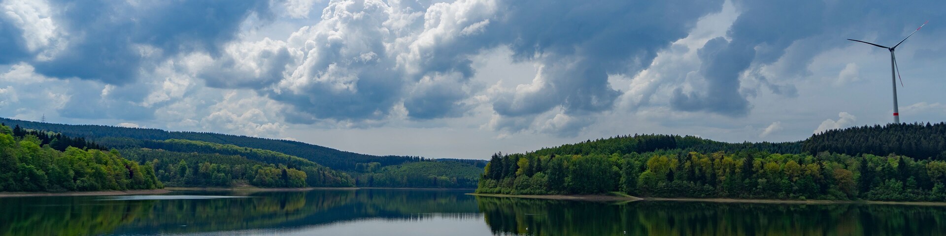Un lago tranquilo con reflejos de nubes y árboles, rodeado de colinas boscosas y una turbina eólica.
