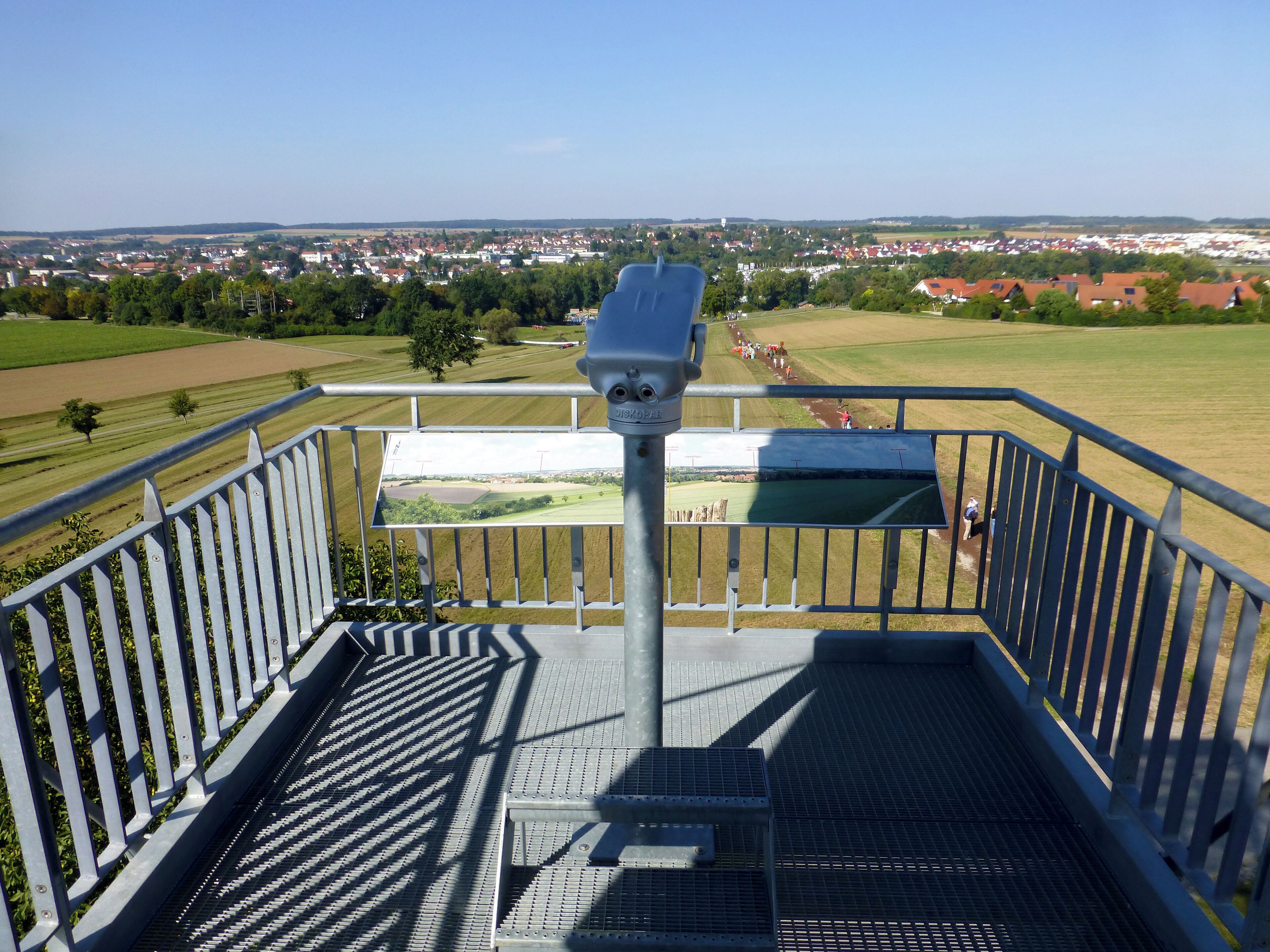A metal observation platform with a telescope, overlooking a rural landscape with fields and scattered houses.