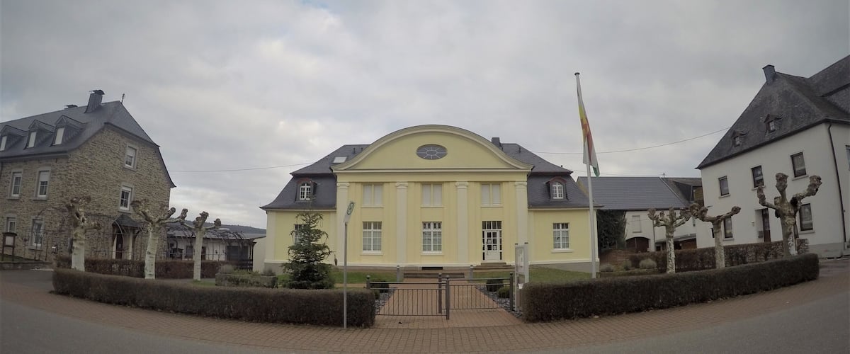 A yellow building with a grey roof, surrounded by stone buildings and trees.