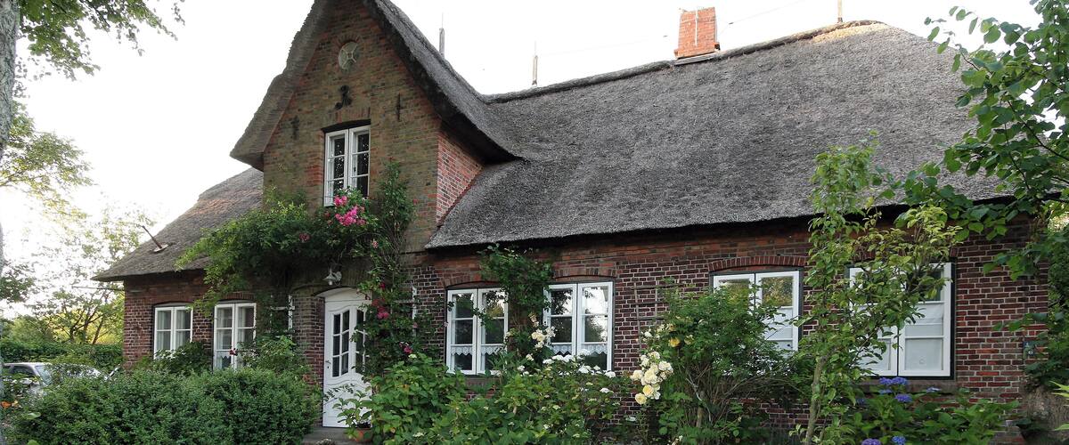A quaint brick house with a thatched roof, surrounded by lush greenery and blooming flowers.