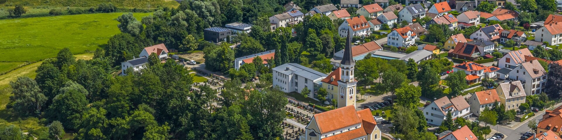 Vista aérea de un pueblo con casas de tejados rojos, una iglesia con un campanario alto y campos verdes circundantes.
