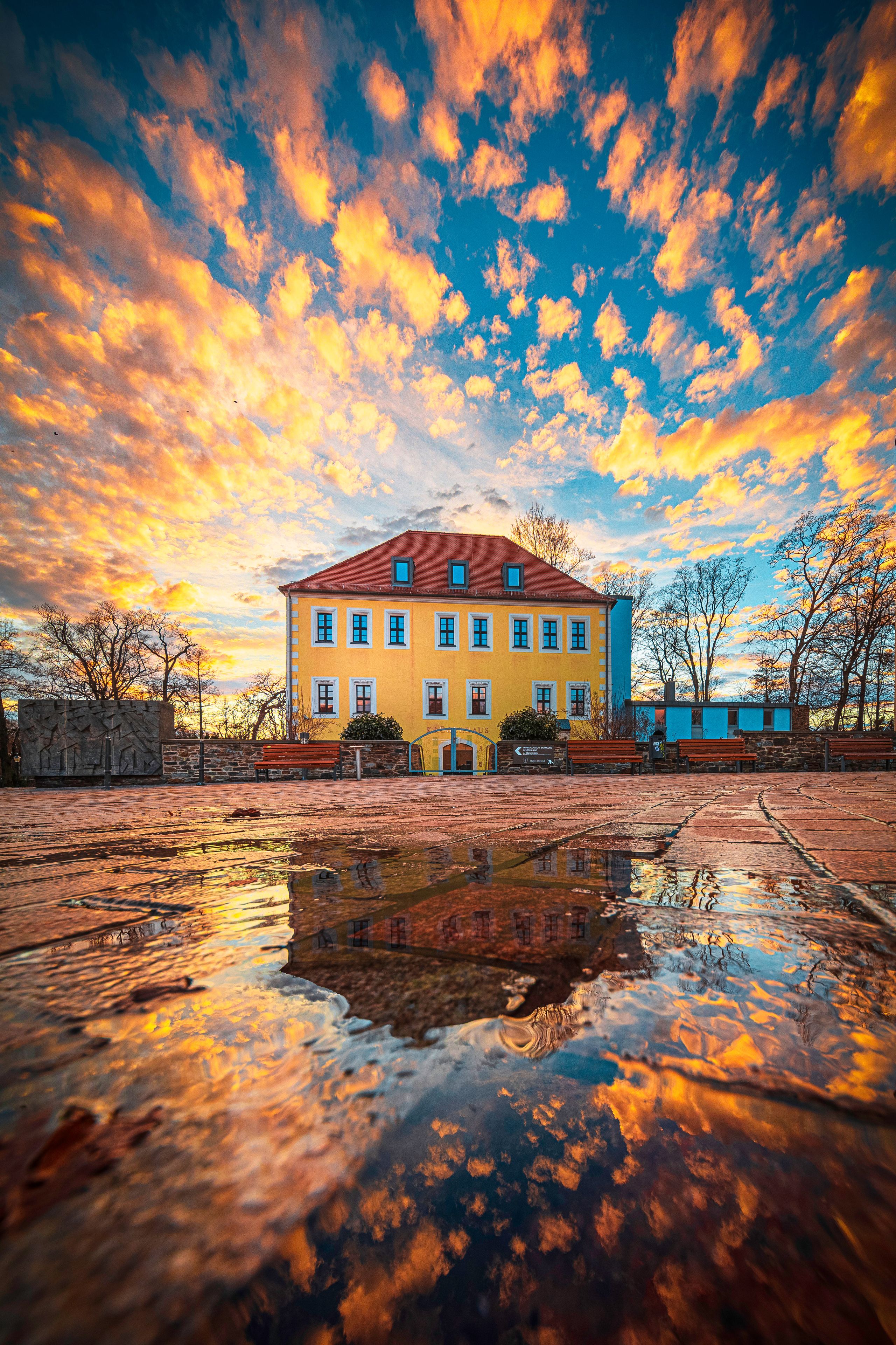 A yellow building with a red roof, surrounded by trees and a wet reflective ground.