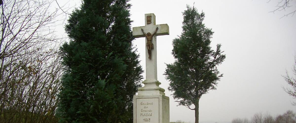 A white cross monument with a crucifix, surrounded by greenery and trees.