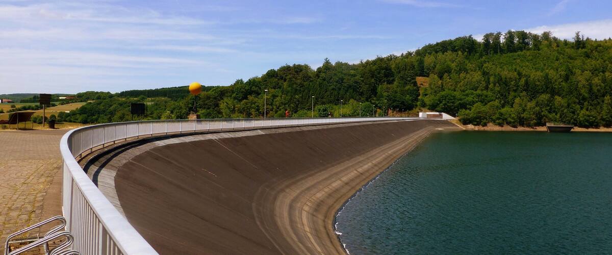 A dam with a curved spillway, a boat on the water, and a forested hillside in the background.