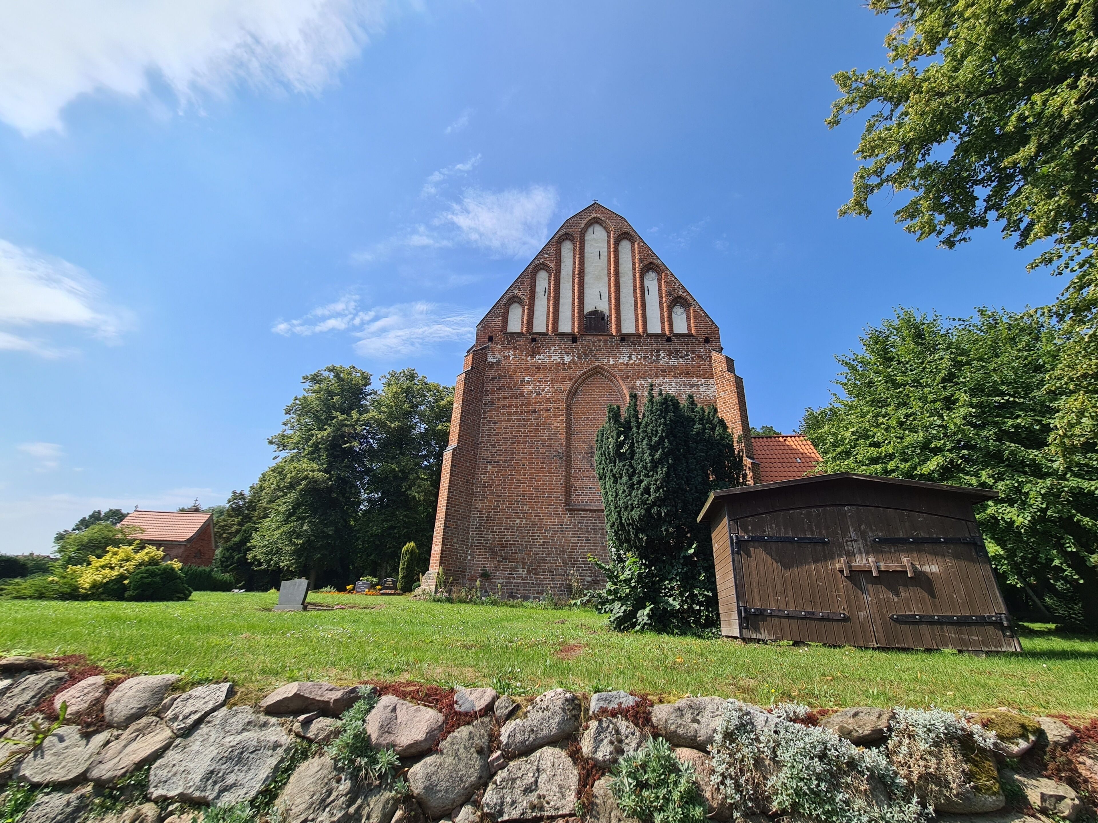 A brick church with a pointed roof and a bell tower, surrounded by a stone wall and greenery.