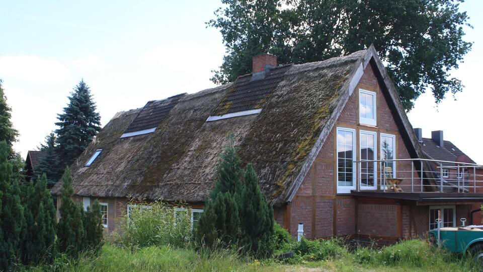 A thatched-roof house with a brick extension, surrounded by greenery and a parked vintage car.