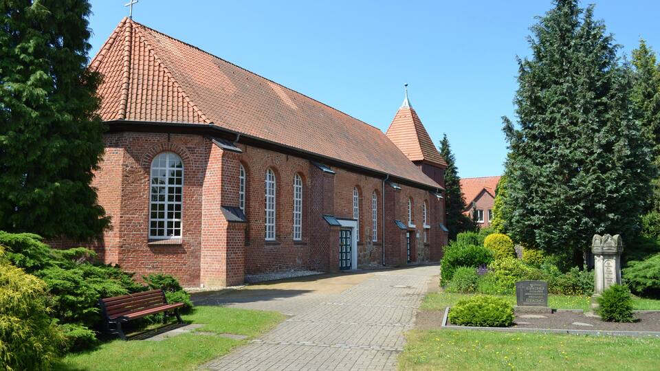 A brick church with a red-tiled roof, a cross on top, and a stone monument in the foreground.