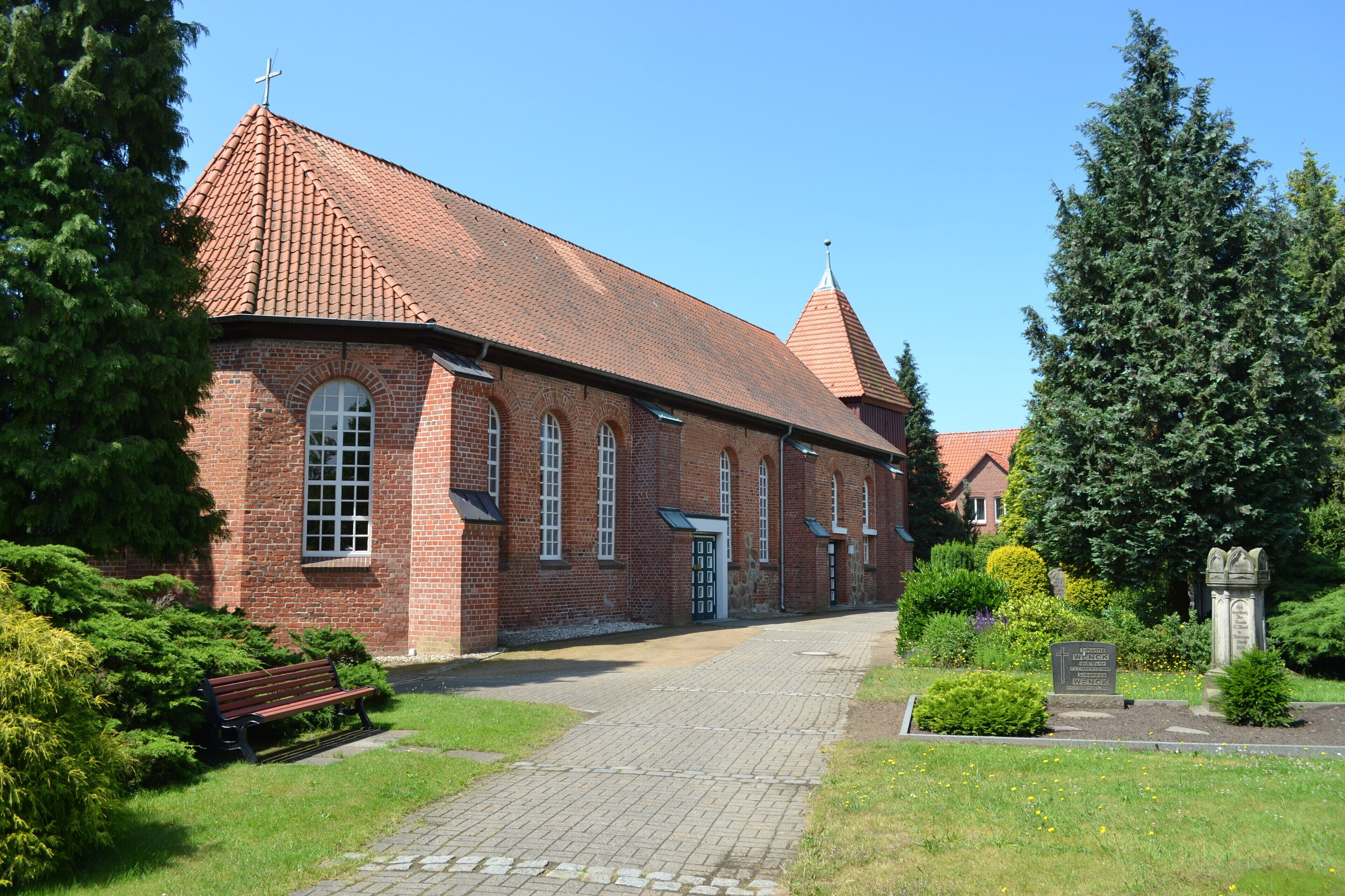 A brick church with a red-tiled roof, a cross on top, and a stone monument in the foreground.