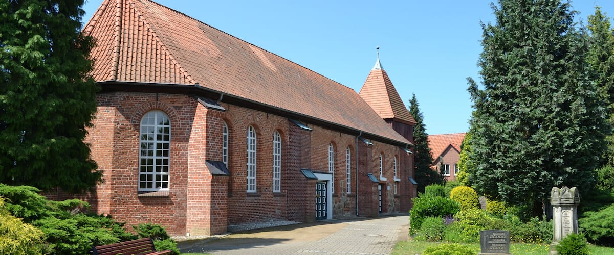 A brick church with a red-tiled roof, a cross on top, and a stone monument in the foreground.