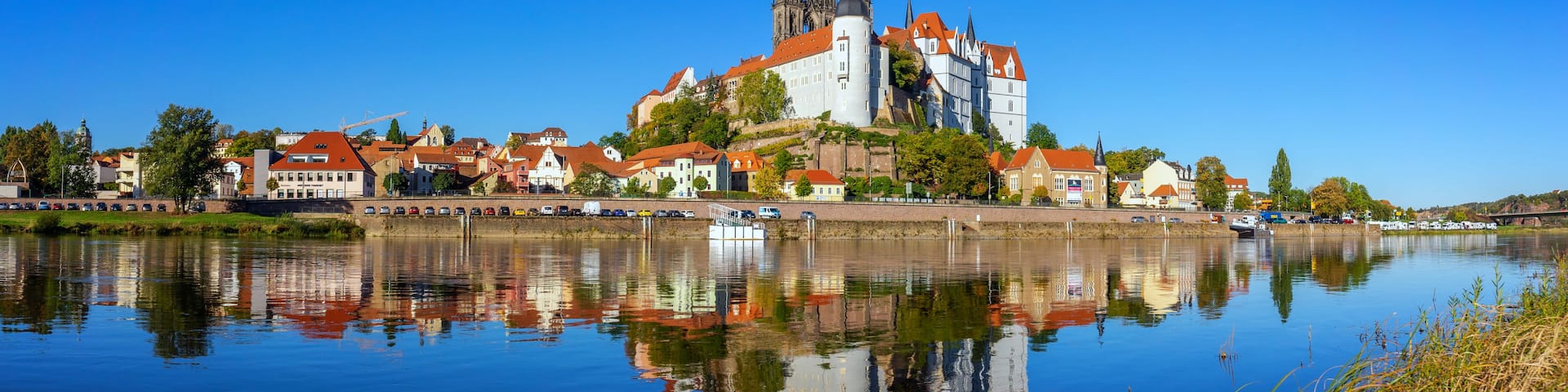 A historic castle with red roofs, a river reflecting its image, and a clear blue sky.