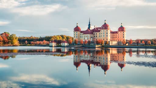 A large palace with red domes and towers, reflecting on a calm lake, surrounded by autumn-colored trees.