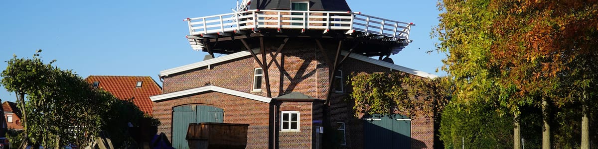 A windmill with a tall tower and a fenced platform, surrounded by trees and a brick building.