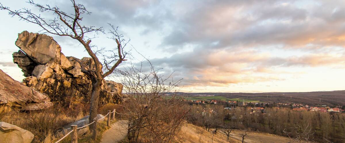 A dirt path with a rope fence, rocky outcrops, and a distant view of a town.