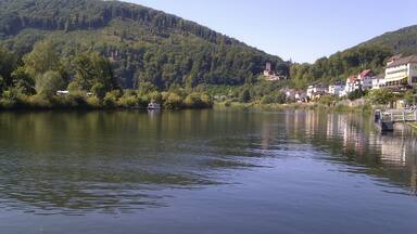 A calm river with a bridge, surrounded by lush green hills and buildings along the riverbank.