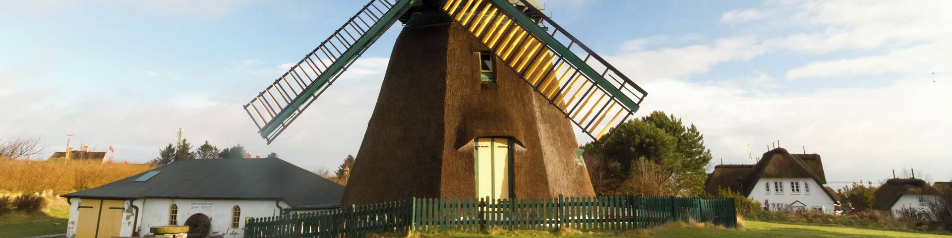 A traditional windmill with a wooden structure and a thatched roof, surrounded by a green fence and a grassy field.