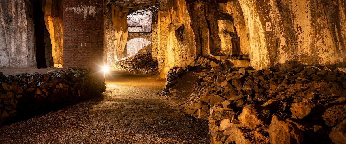 A dimly lit cave with a narrow passageway, stone walls, and a large rock formation in the distance.