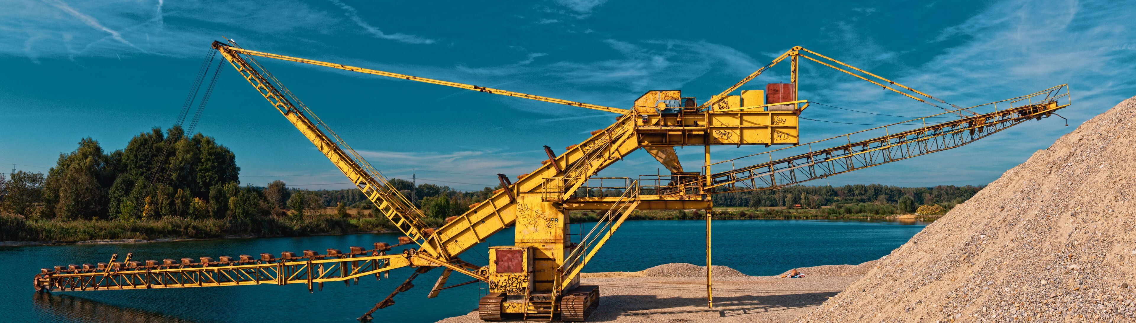 A large yellow industrial crane by a river with a pile of gravel and a forest in the background.