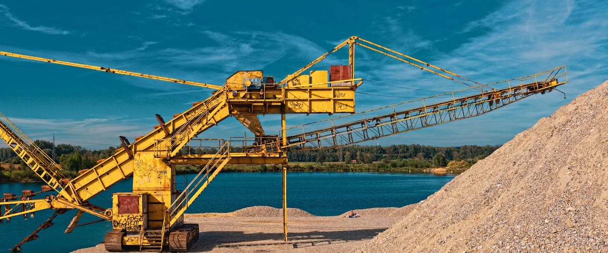 A large yellow industrial crane by a river with a pile of gravel and a forest in the background.