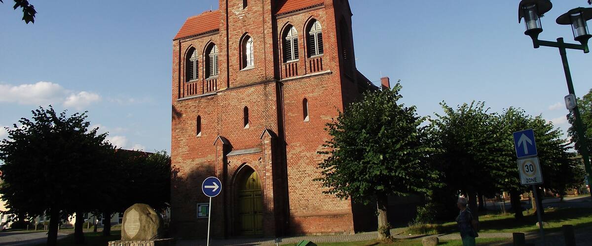 A large brick church with a tall tower and a clock. There are trees and a road with a speed limit sign.