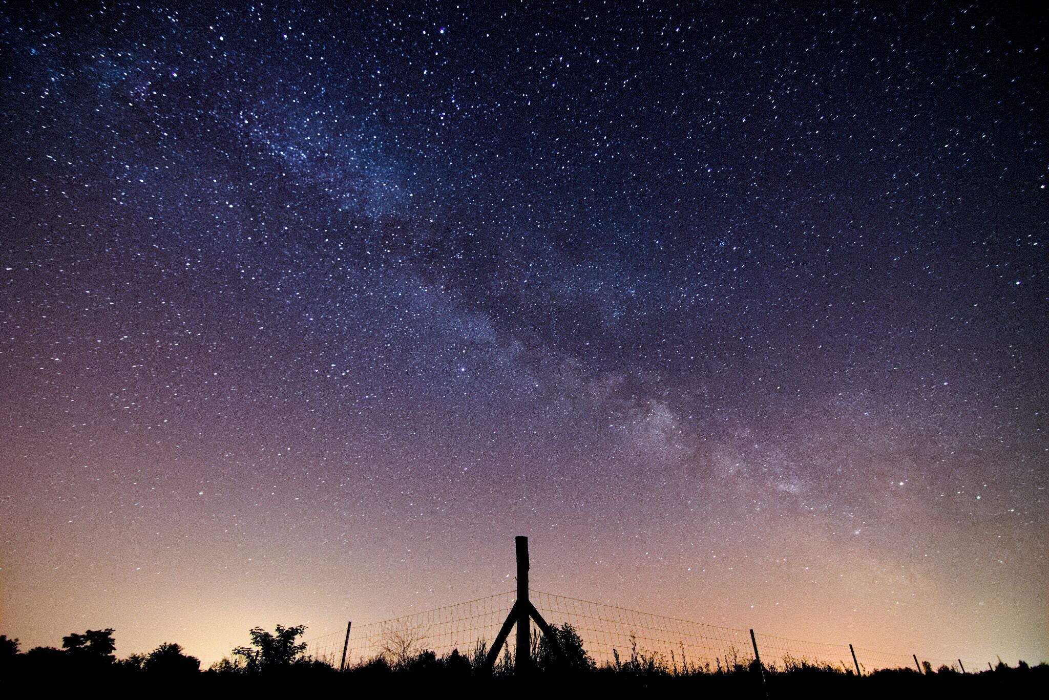A starry night sky with the Milky Way visible, a fence, and silhouettes of trees.