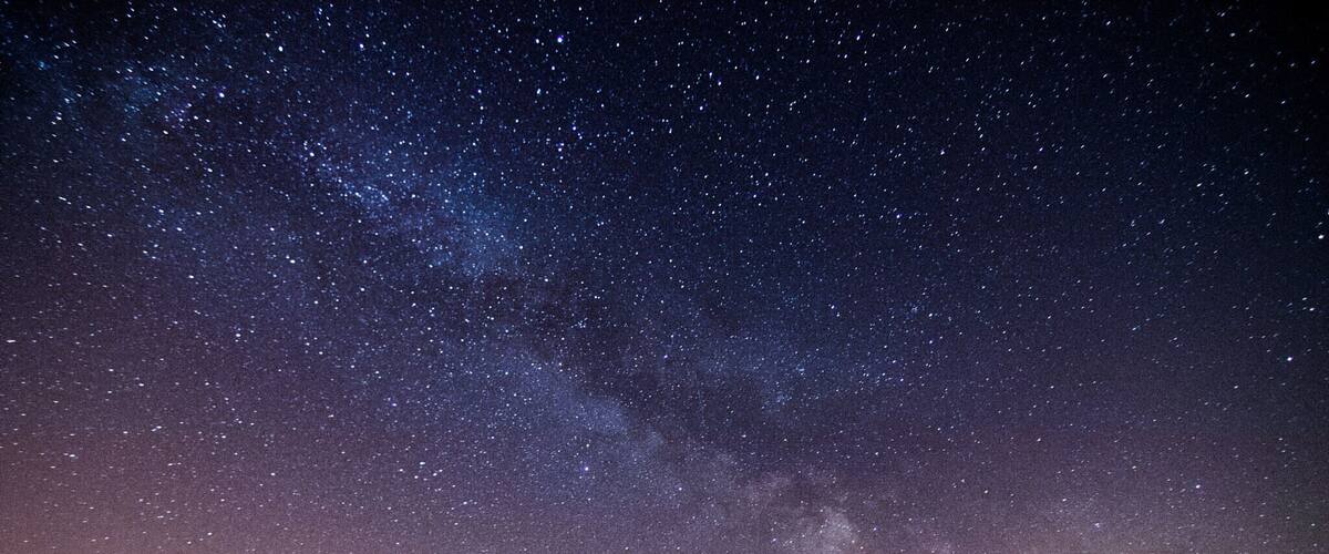 A starry night sky with the Milky Way visible, a fence, and silhouettes of trees.