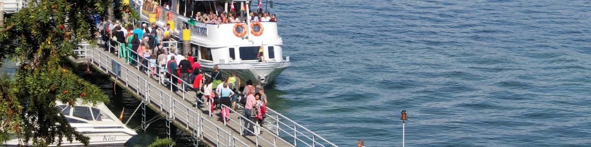 Une jetée où des gens se promènent, un bateau transportant des passagers et, au loin, une côte bordée de bâtiments.