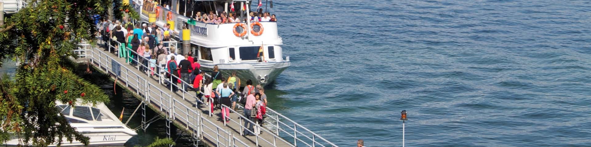 A pier with people walking, a boat with passengers, and a distant shoreline with buildings.