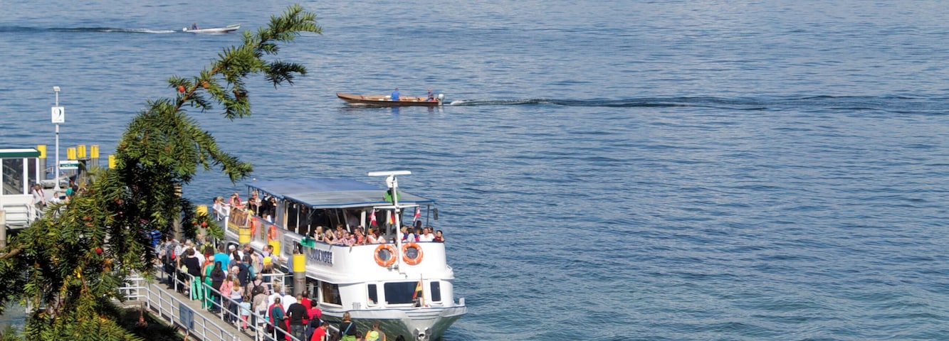 Un muelle con gente caminando, un barco con pasajeros y una costa distante con edificios.