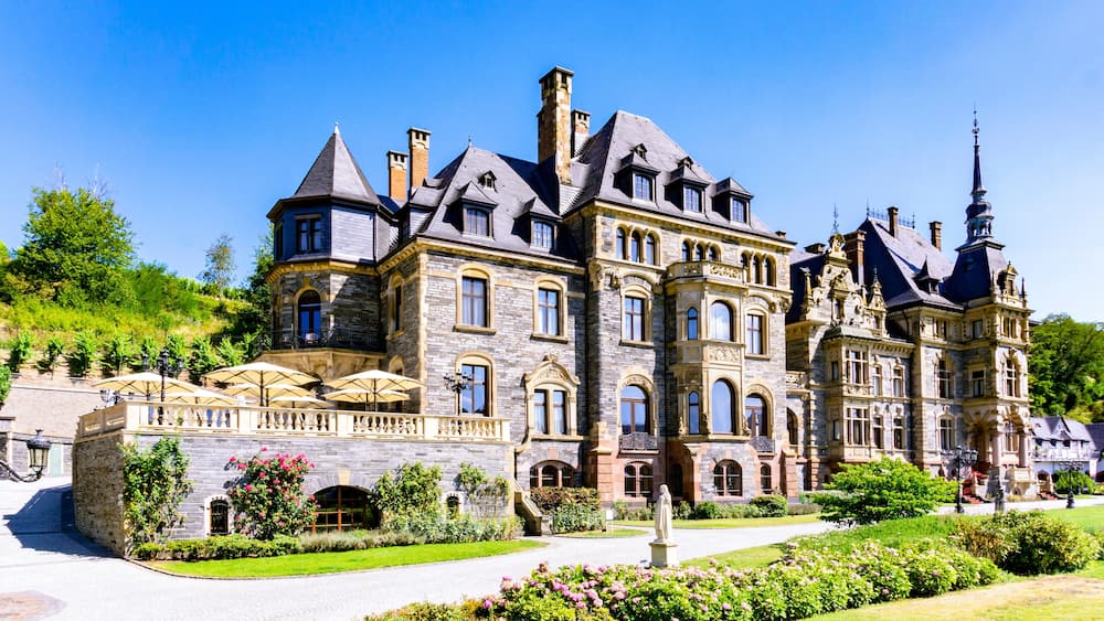Un grand bâtiment historique en pierre, doté de nombreuses tours et tourelles, entouré de jardins luxuriants et bordé d’un chemin pavé.