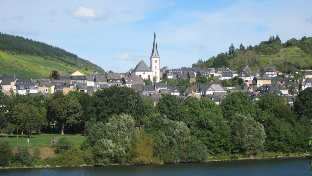 Une ville dont l’église, reconnaissable à son clocher imposant, est entourée de maisons et d’une végétation luxuriante.